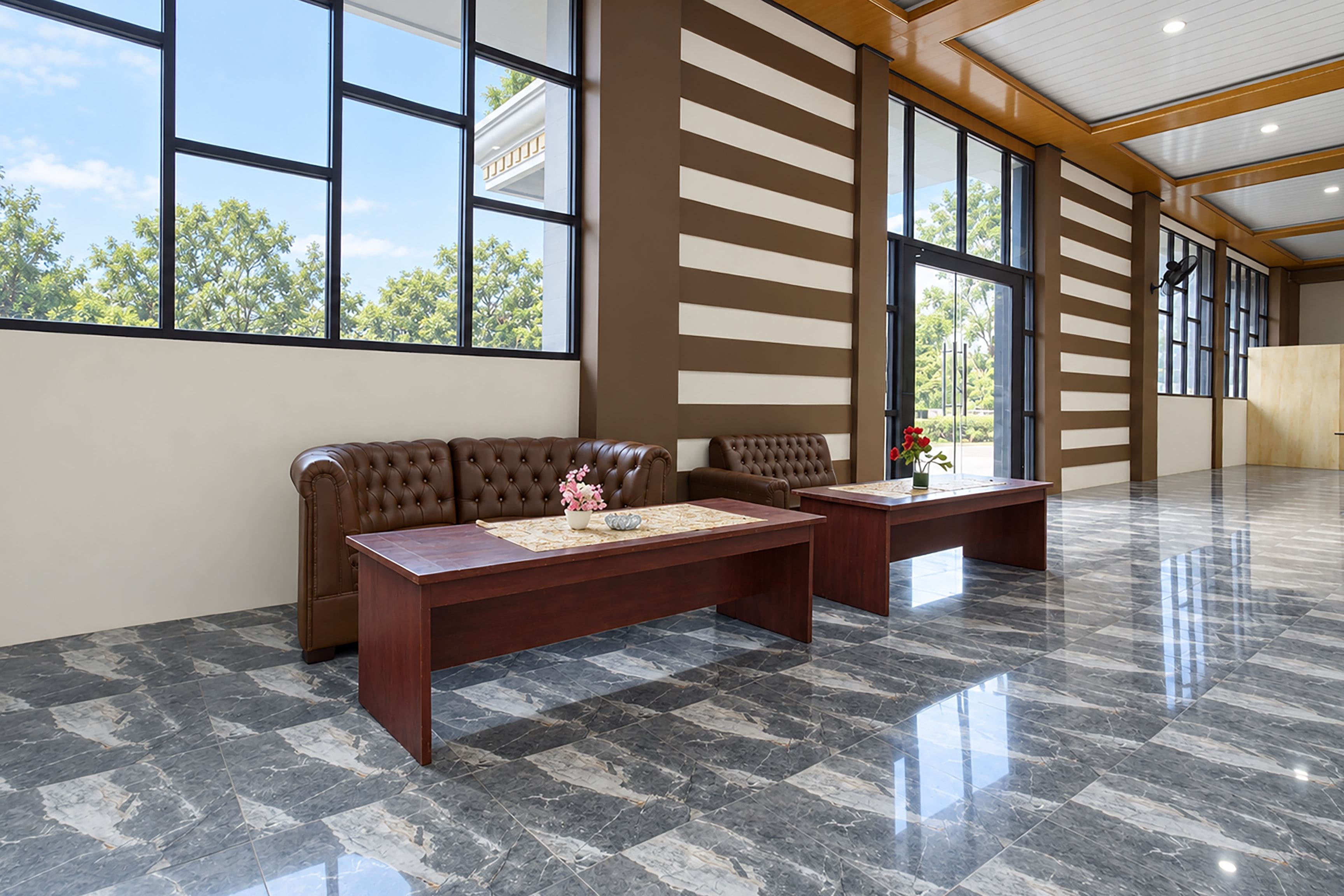 A spacious room with a brown leather sofa, a wooden coffee table, and a desk with a vase of flowers. Large windows allow natural light to enter.