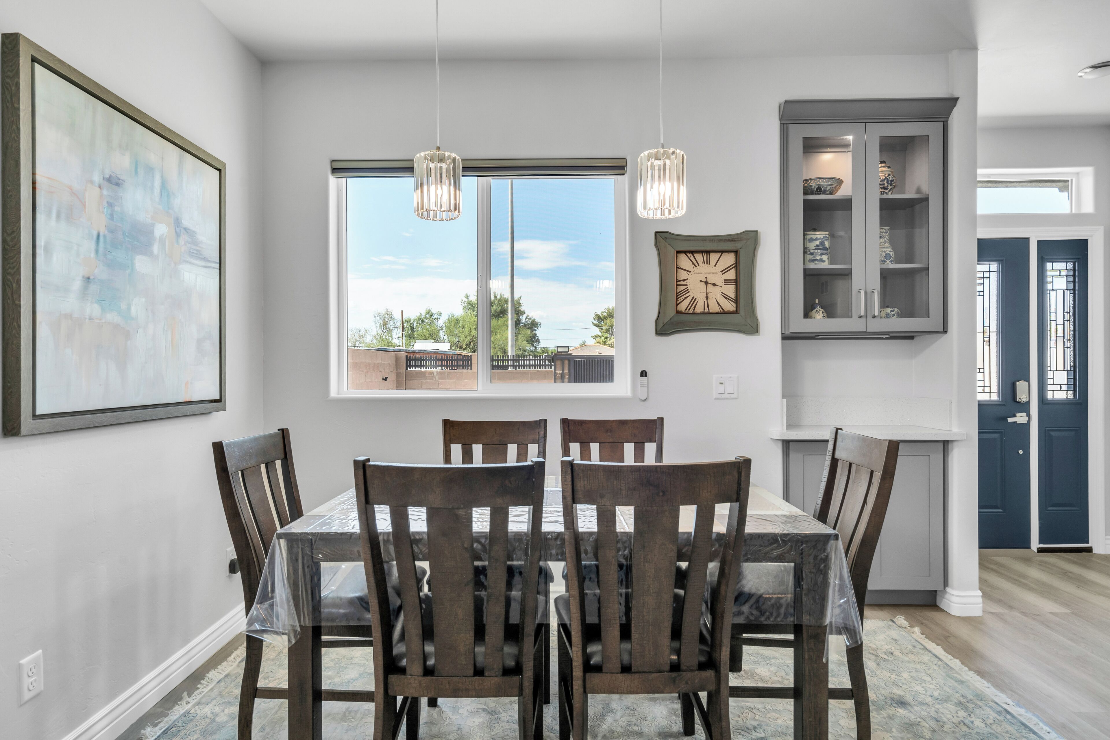 A dining area with a table and chairs, a window with a view, a clock, and a cabinet with decorative items.