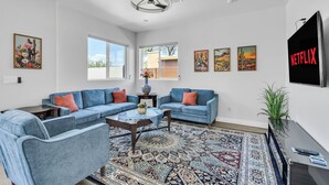 A living room with a sectional sofa, a coffee table, and a patterned rug. There are windows, artwork on the walls, and a television.