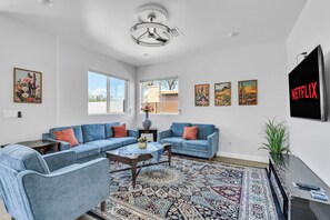 A living room with a sectional sofa, a coffee table, and a patterned rug. There are windows, artwork on the walls, and a television.