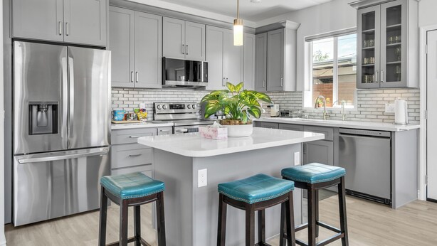 A modern kitchen with grey cabinets, stainless steel appliances, a central island with stools, and a window providing natural light.