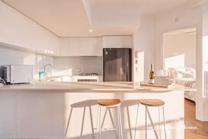 A modern kitchen with white cabinetry, a stainless steel refrigerator, a built-in gas stove, and a large island with bar stools. - "SUNSET" By TheGuestHouse. (Palm Beach)