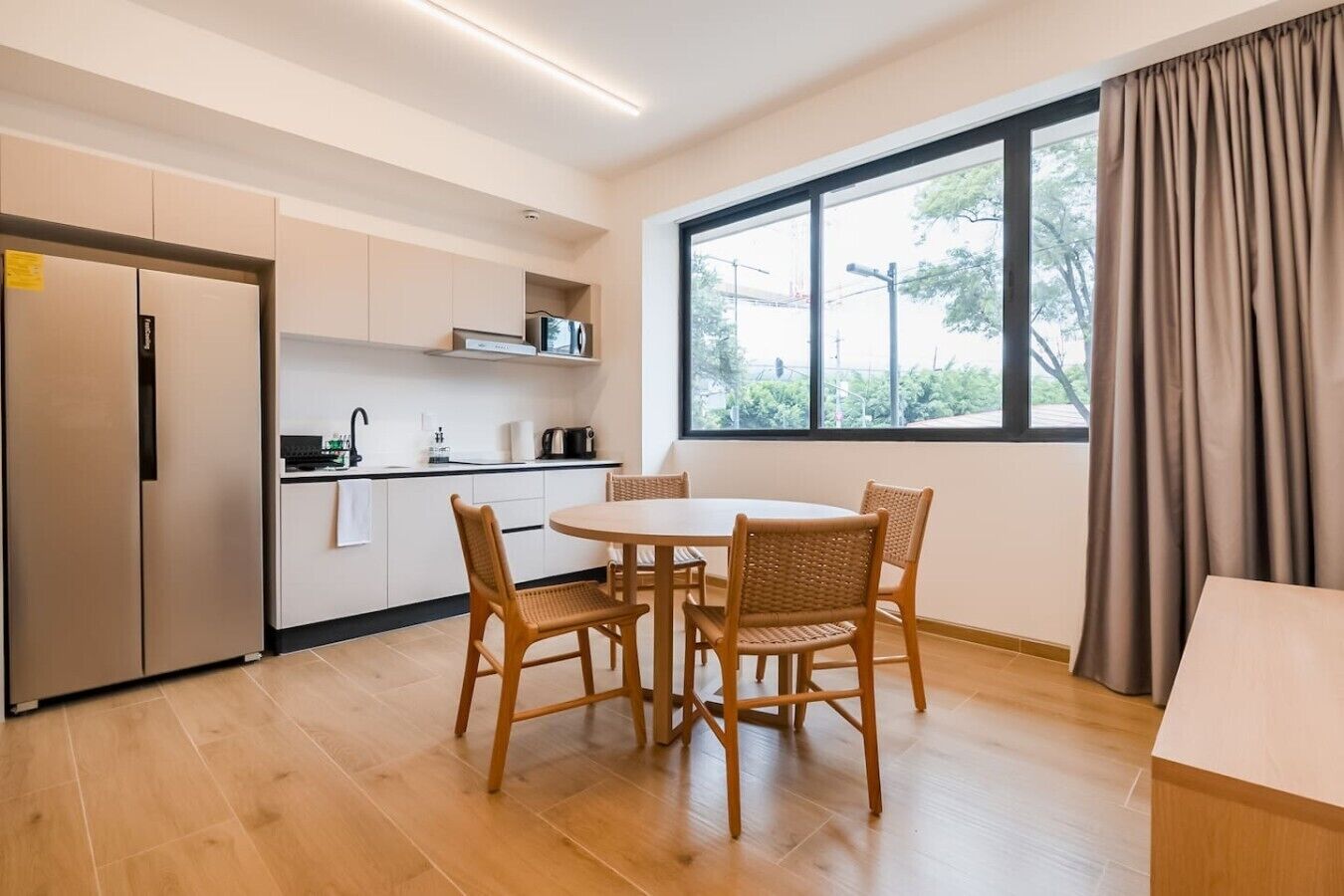 A modern kitchen with a dining table and chairs, a refrigerator, and a large window with curtains.
