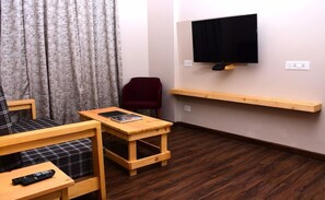 Quarto pequeno com sofá, uma mesa de madeira, uma cadeira vermelha, TV fixada na parede e prateleira de madeira abaixo da TV.