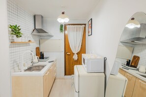 A compact kitchen with a microwave, sink, and a small refrigerator. There is a wooden door with a curtain, a hanging plant, and a shelf with wine glasses.