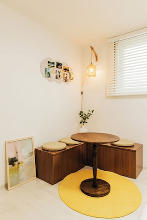 A room with a round wooden table, a yellow woven rug, a wall-mounted light fixture, and a bulletin board with photos and notes.