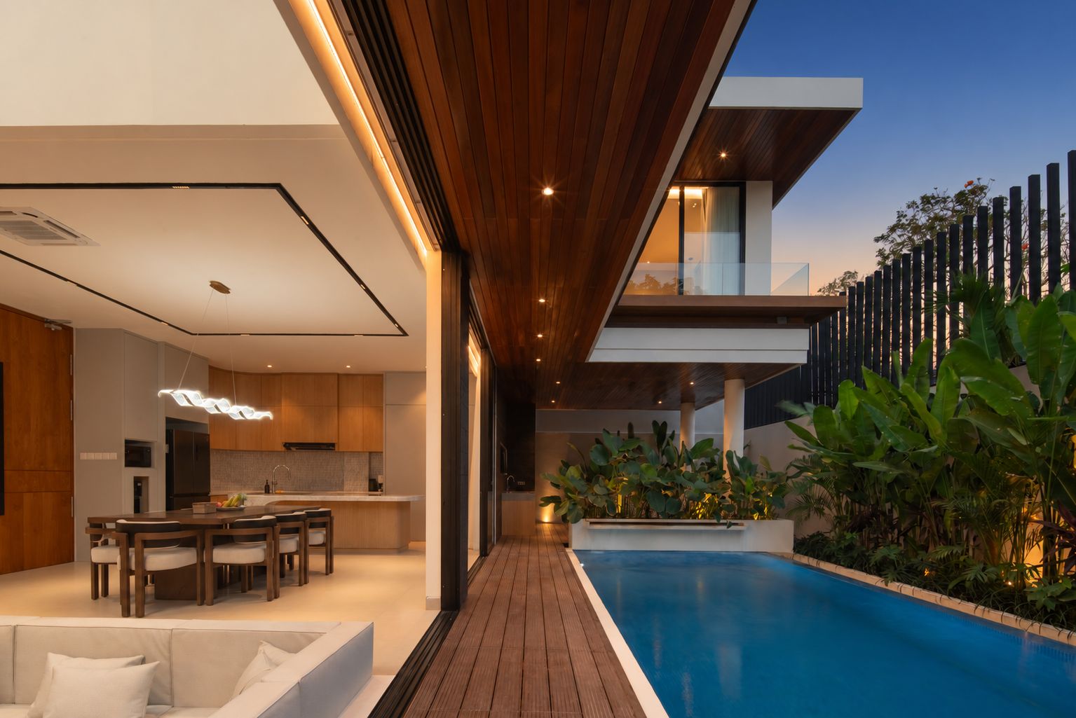 A modern outdoor pool area with a wooden deck, a dining area with a table and chairs, and a kitchen visible in the background.