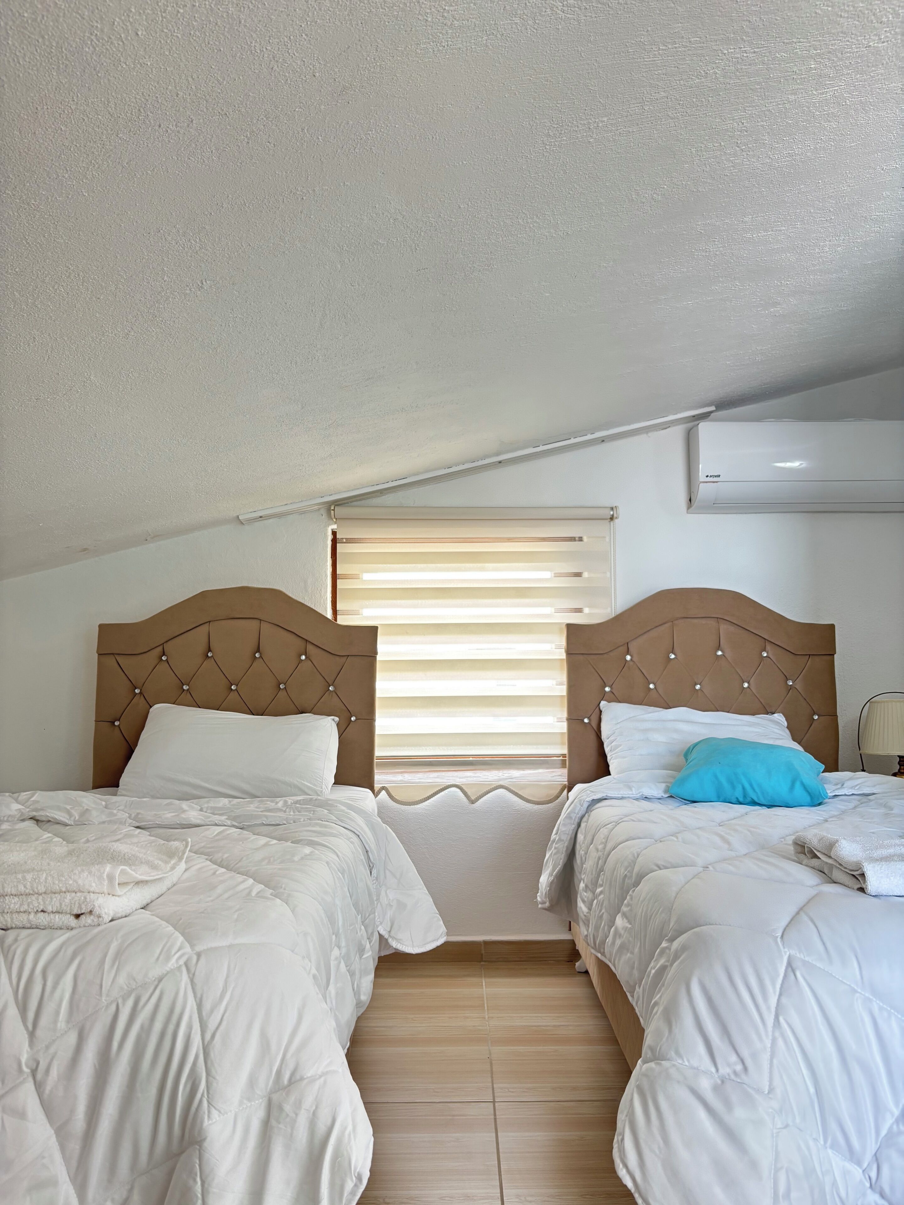 Ein Schlafzimmer mit zwei Betten, einem Fenster mit Jalousien, einer Klimaanlage und einer Lampe auf dem Nachttisch.