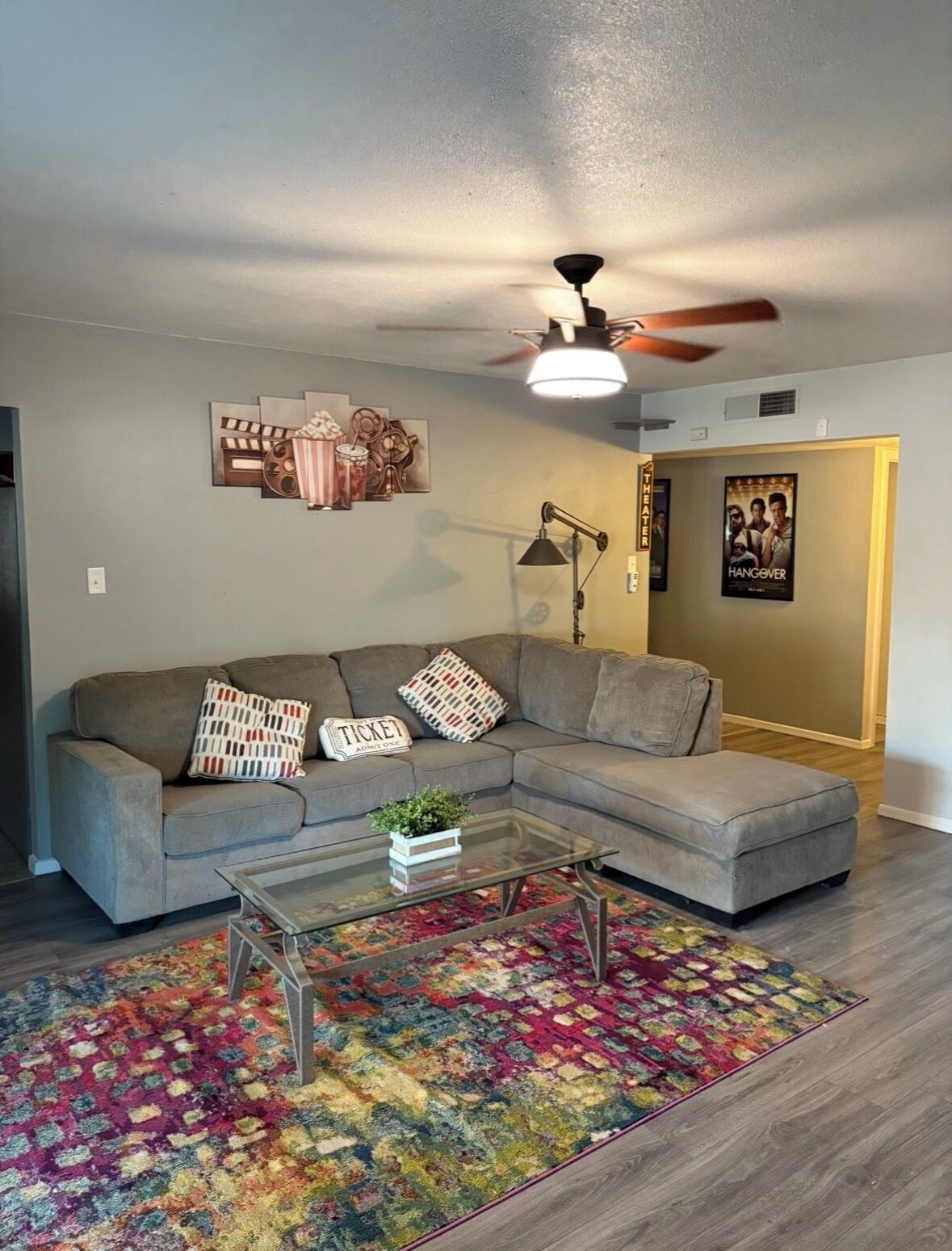 A living room with a sectional sofa, a glass coffee table, a colorful rug, a ceiling fan, and a poster on the wall.