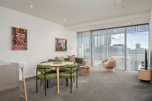 A modern living room with a round table, green chairs, a sofa, and a large window with a city view. - Downtown Stylish Apartment w/ Pool, Gym & Parking! (West Melbourne)