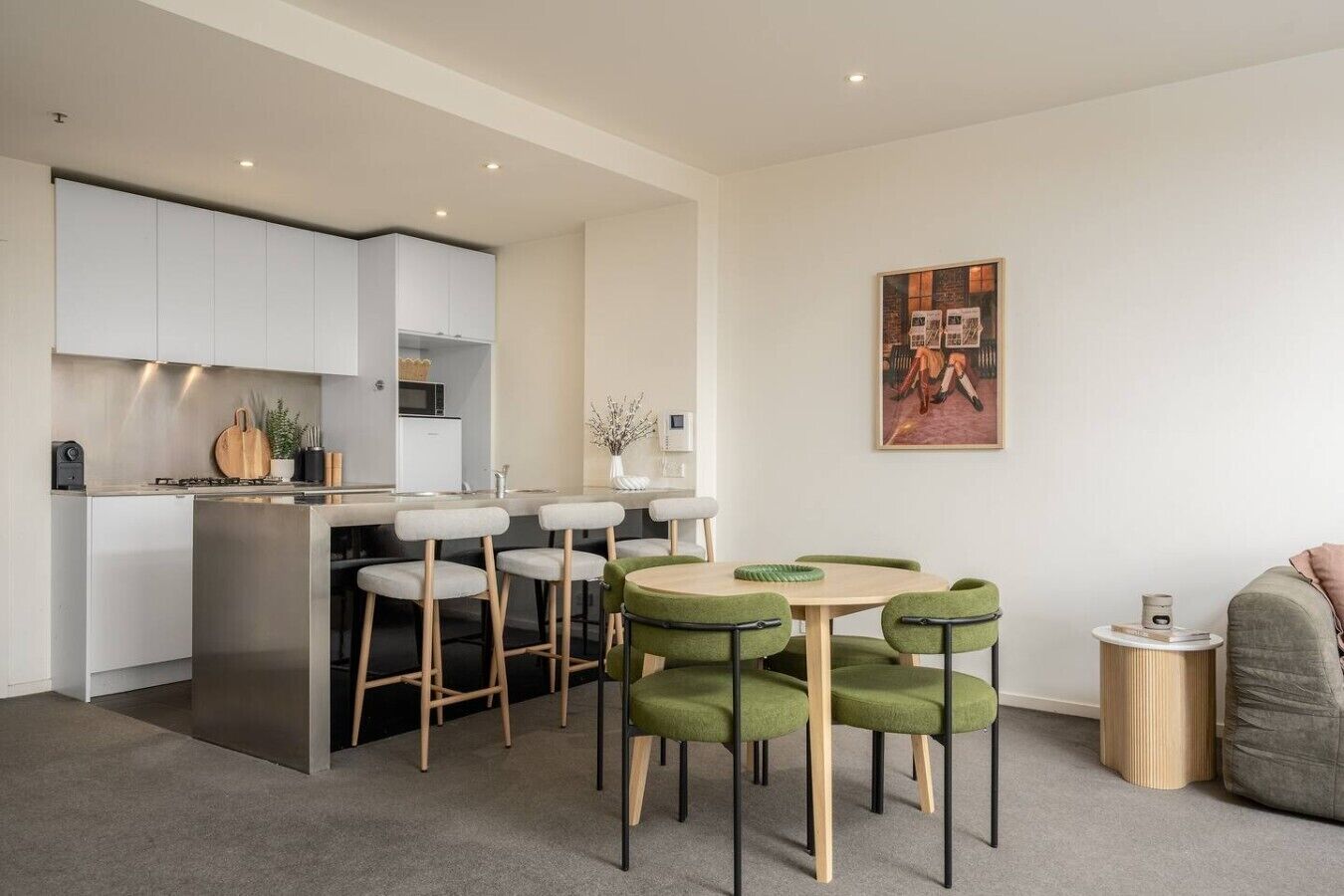 Une cuisine moderne avec des meubles blancs, un îlot central doté de tabourets et un coin repas avec des chaises recouvertes d’un tissu vert.