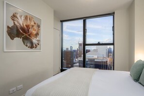 A hotel room with a large bed, a framed artwork on the wall, and a city view through a large window. - Skyscraper CBD Apartment with Pool, Gym & Parking! (Melbourne)