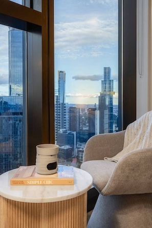 A modern room with a large window, a round wooden side table, a beige armchair, and a book titled 'Simple Chic'. - Skyscraper CBD Apartment with Pool, Gym & Parking! (Melbourne)
