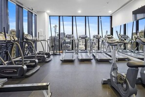 A gym with treadmills, ellipticals, and weight machines, featuring large windows with a cityscape view. - Skyscraper CBD Apartment with Pool, Gym & Parking! (Melbourne)