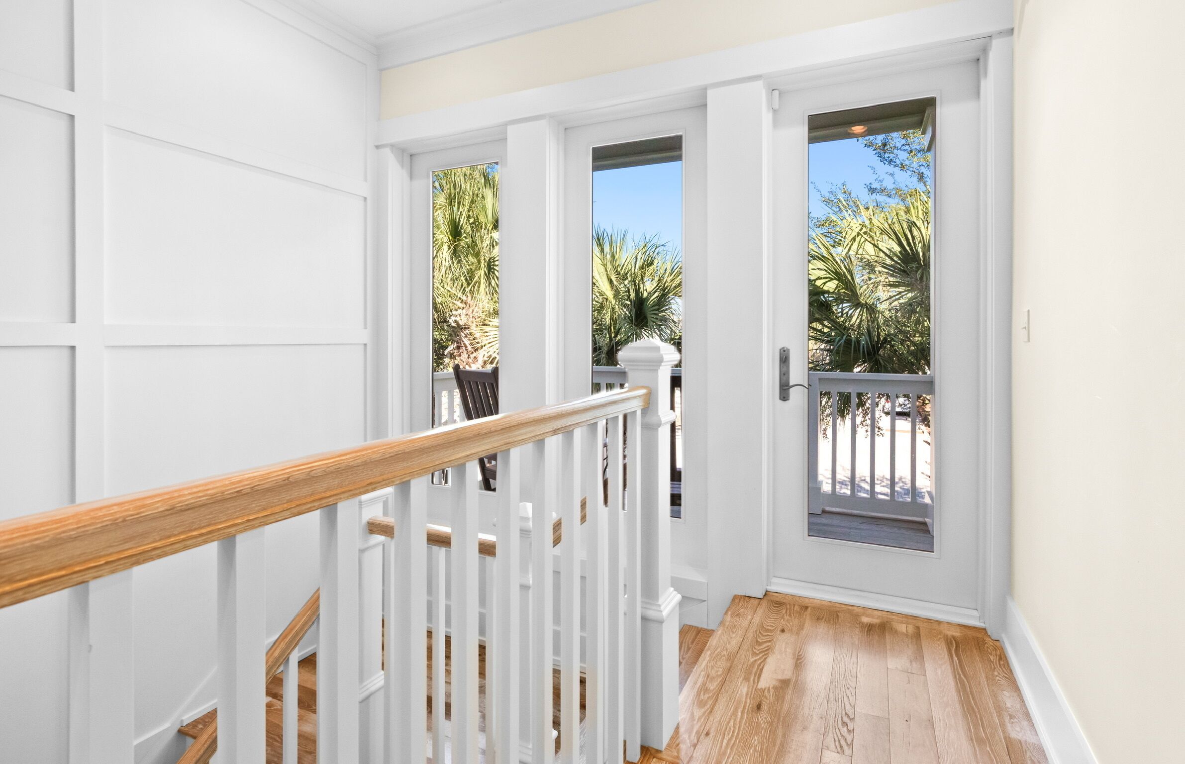 Un couloir intérieur aux murs blancs, avec un escalier en bois menant à une porte donnant sur une vue de palmiers.