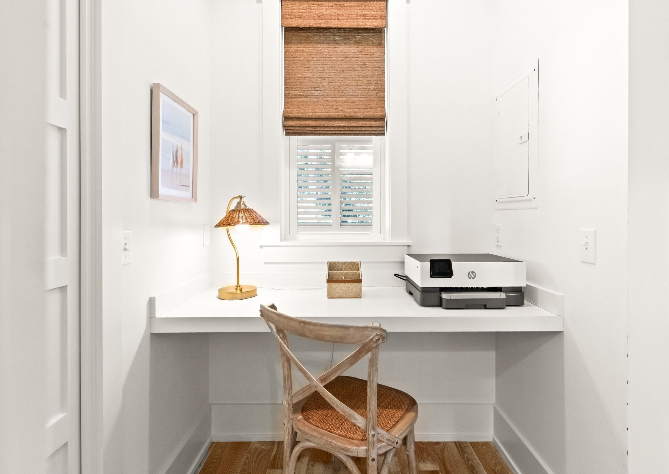 A white desk with a lamp, a printer, and a chair in a room with white walls and a window with blinds.