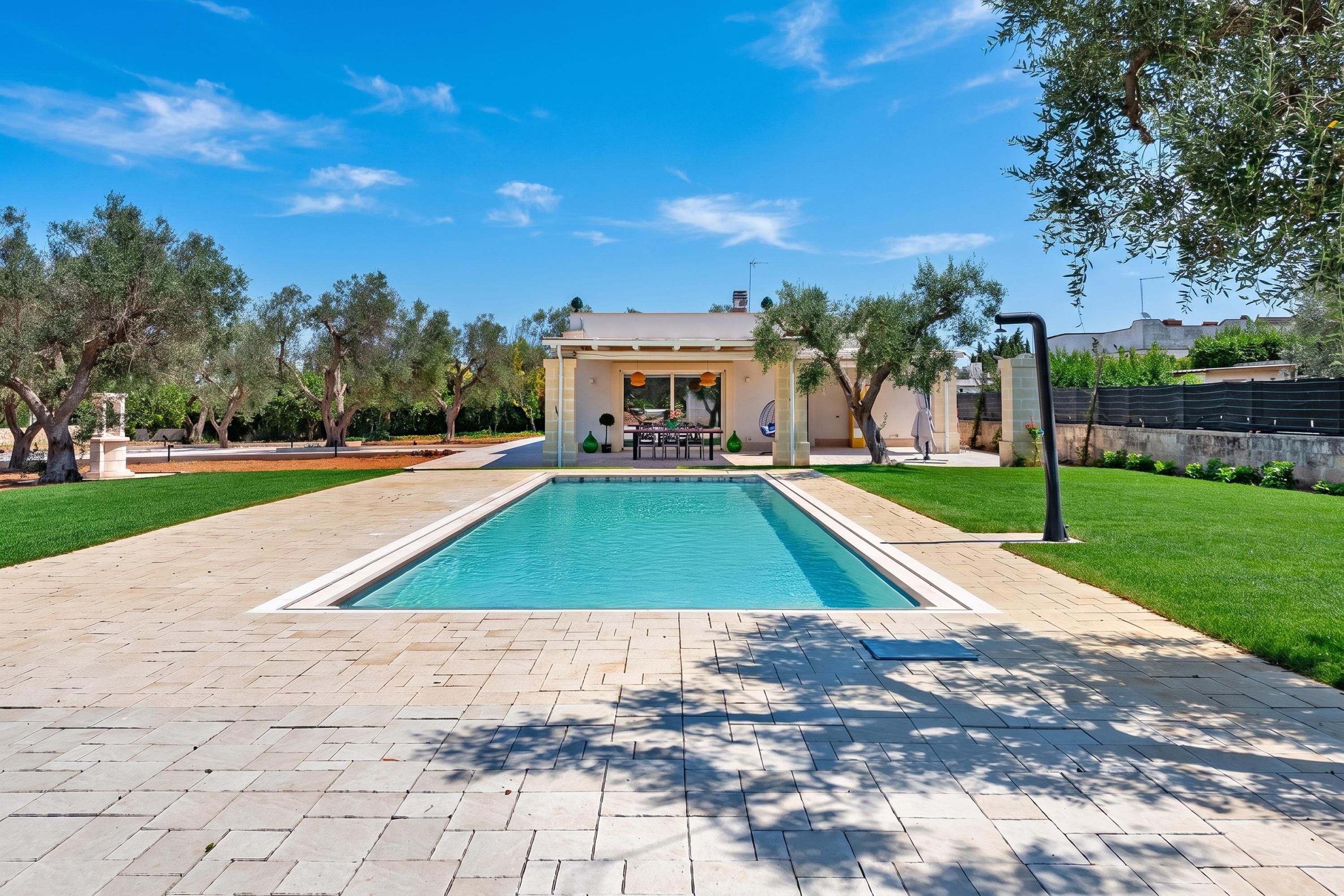 Une piscine rectangulaire avec une terrasse carrelée, entourée d’une pelouse bien entretenue et d’arbres.