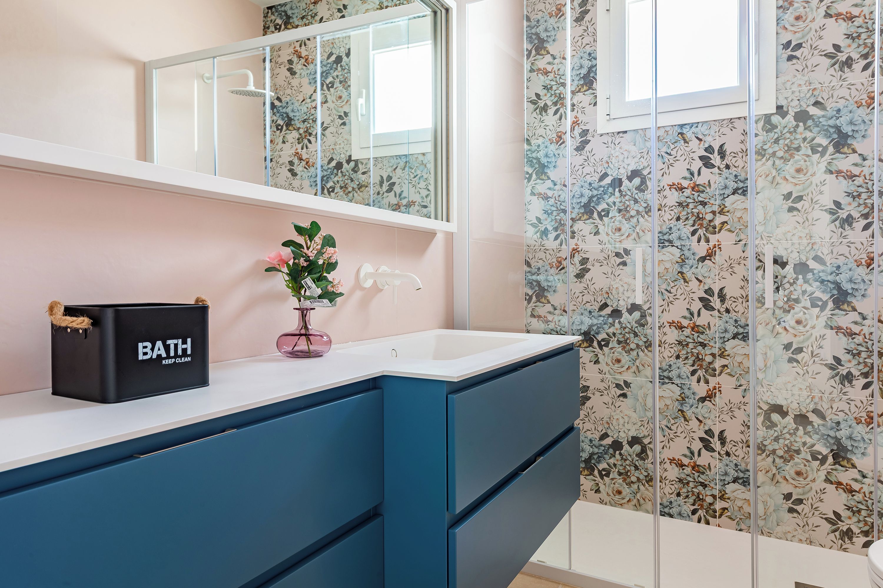 Une salle de bain avec un lavabo blanc, des meubles bleus, un sac de bain noir et un papier peint à motifs floraux.