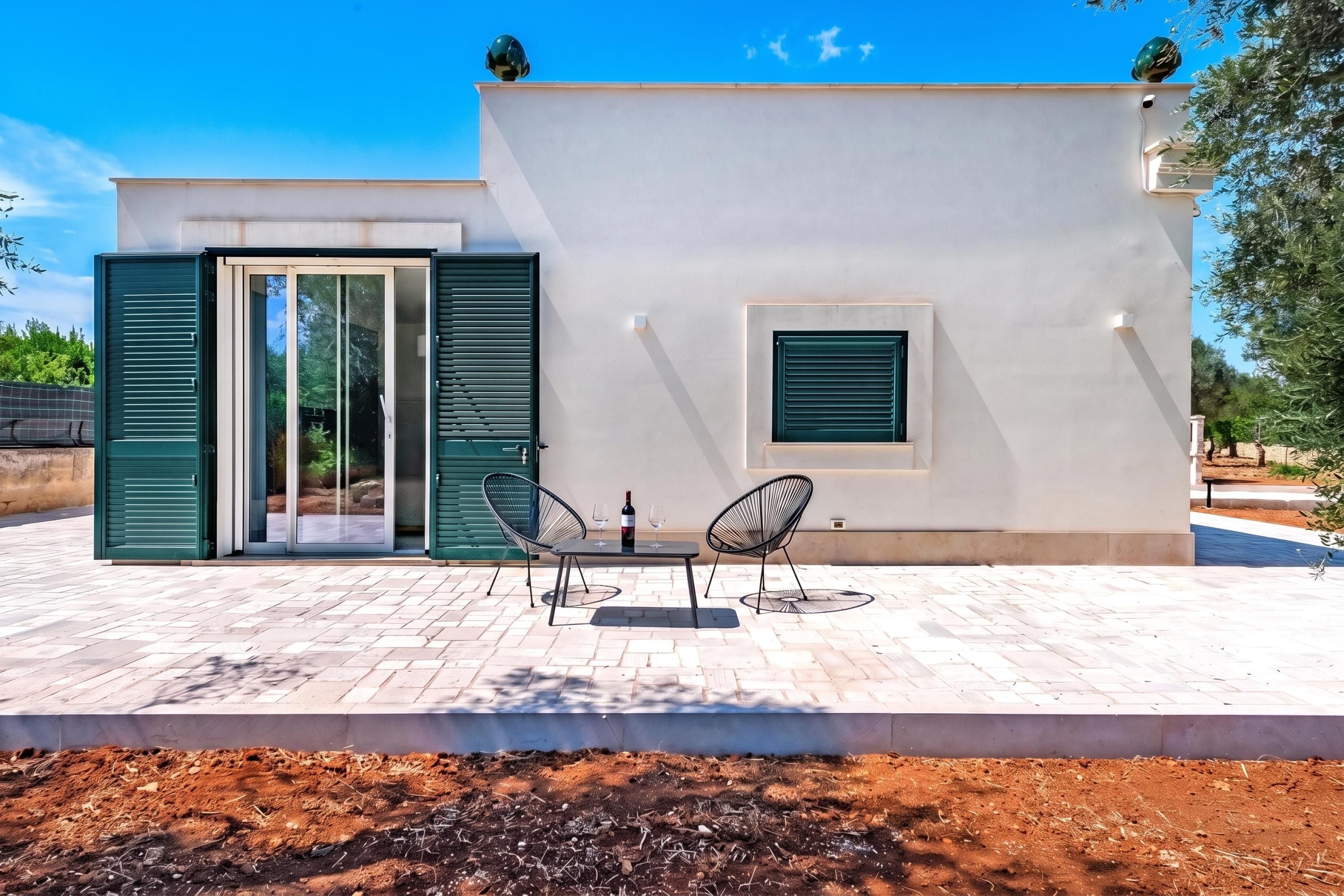 Un edificio moderno con un patio amueblado con dos sillas y una mesa, un gran ventanal corredero y un ventanuilla con contraventanas.