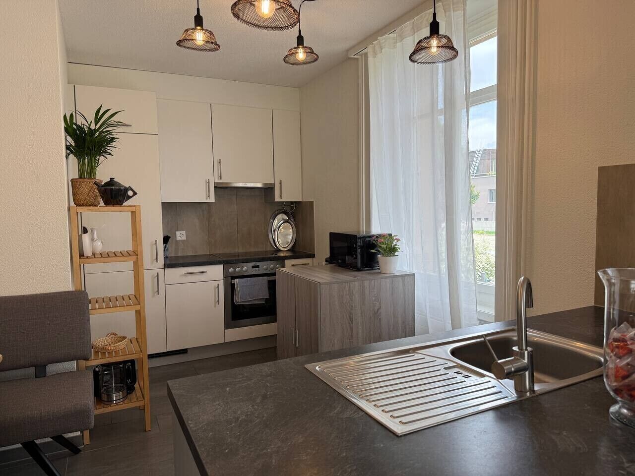 Moderni keittiö, jossa on valkoiset kaapit, sisäänrakennettu uuni ja mikroaaltouuni. Keittiössä on myös puinen hylly, jossa on keittiötarvikkeita, sekä ruukkukasvi.