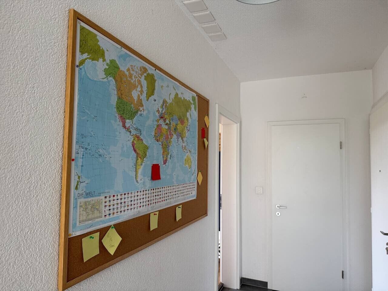 Ein Zimmer mit einer großen Weltkarte an der Wand, einer Tür und einem Korkbrett mit Notizen.