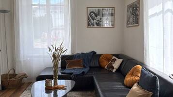Ett modernt vardagsrum med en svart lädersoffa, ett soffbord i glas och en mönstrad matta. På väggen hänger två inramade tavlor och i rummet står en golvlampa.