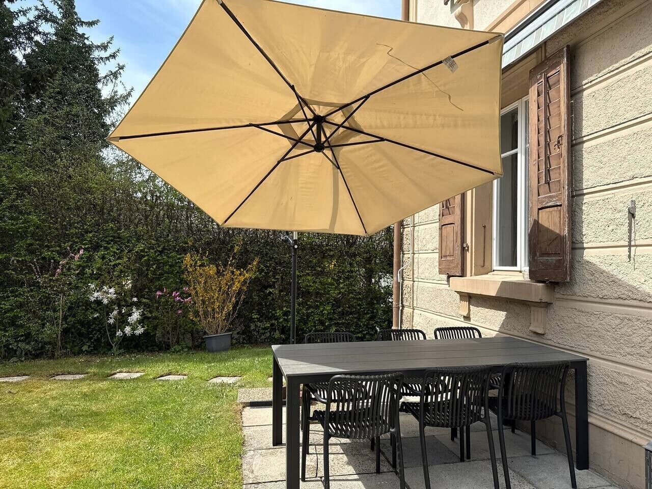 Eine Außenpatio mit Tisch und Stühlen, einem großen Sonnenschirm und einem Gebäude mit Fenstern und Fensterläden im Hintergrund.