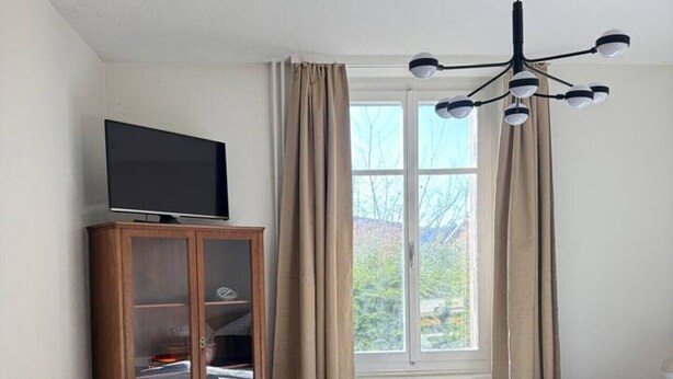 A bedroom with a bed, a television on a wooden cabinet, and a window with curtains. - Spacious apartment with a private garden and entrance (Huttwil)