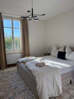 Ein Schlafzimmer mit einem Bett, einem Nachttisch, einer Lampe und einem Fenster mit Vorhängen.