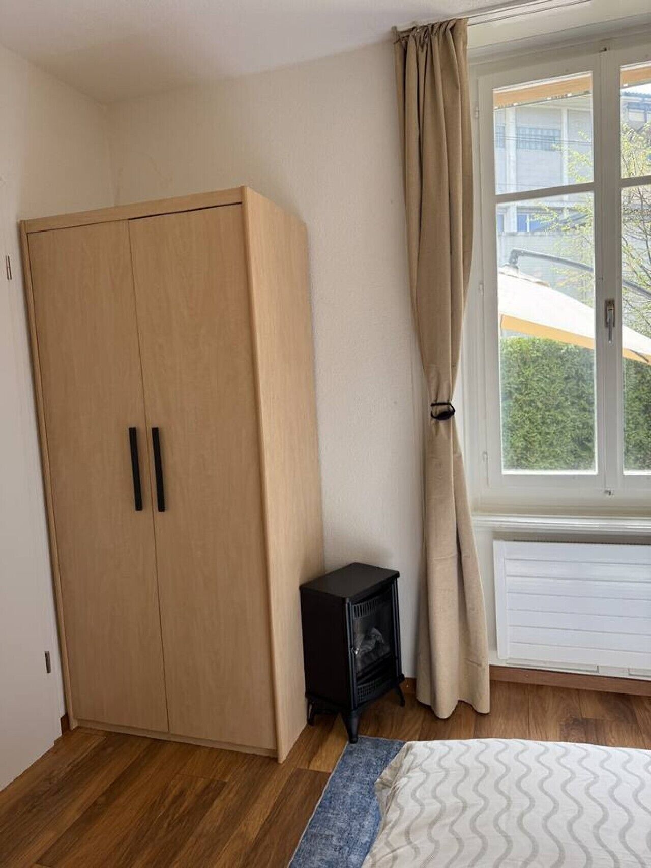 A room with a wooden wardrobe, a small black heater, a window with curtains, and a bed with a patterned bedspread.