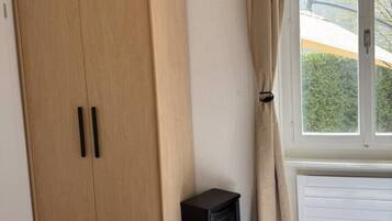 A room with a wooden wardrobe, a small black heater, a window with curtains, and a bed with a patterned bedspread.