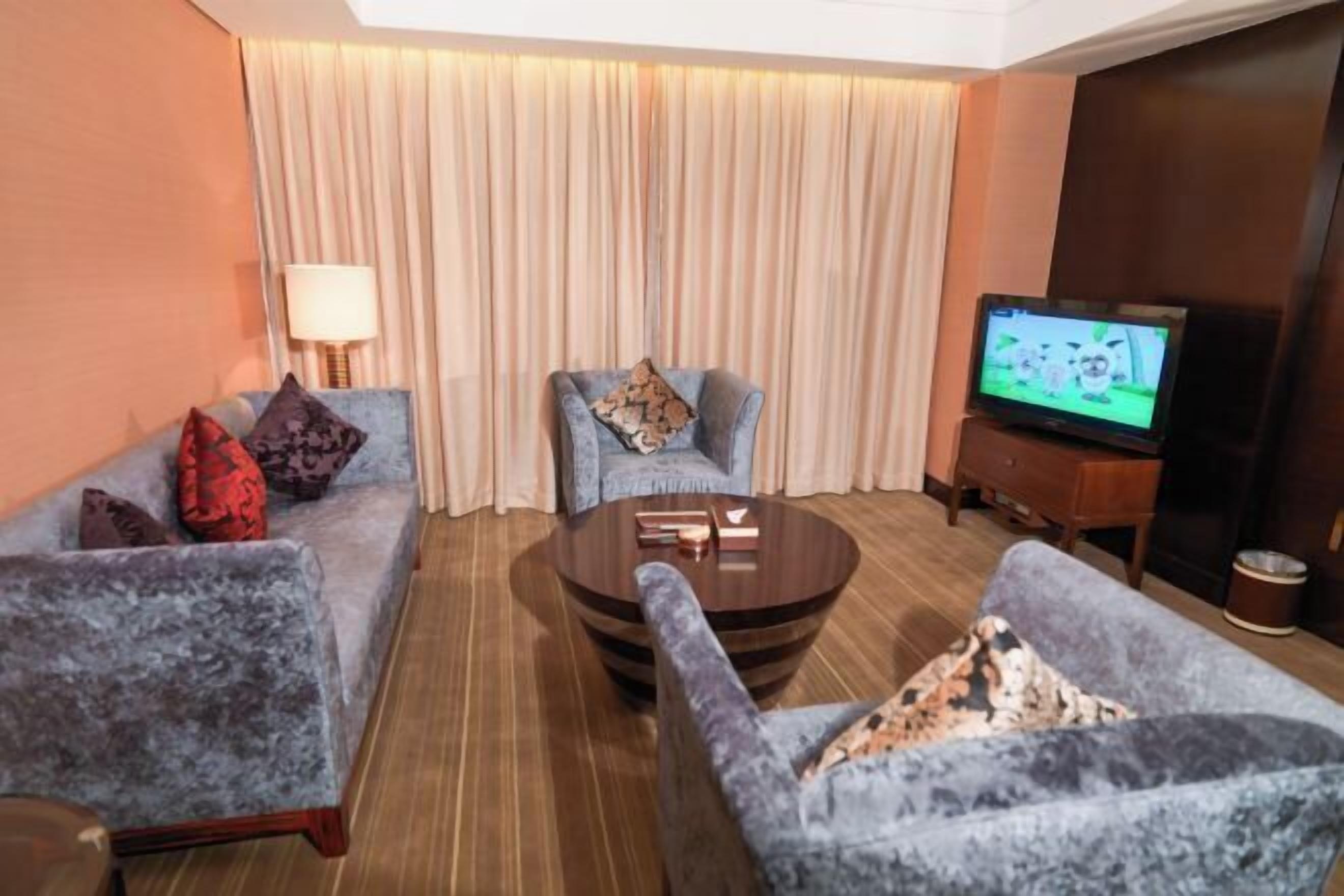 Habitación de hotel con sofá, sillones, una mesa de centro, televisor y cortina.