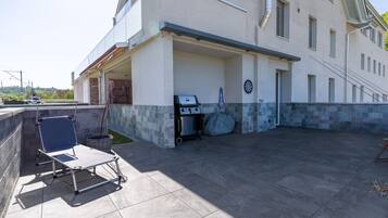 Eine überdachte Terrasse im Freien mit Grill, Klappstuhl und kleinem Tisch.