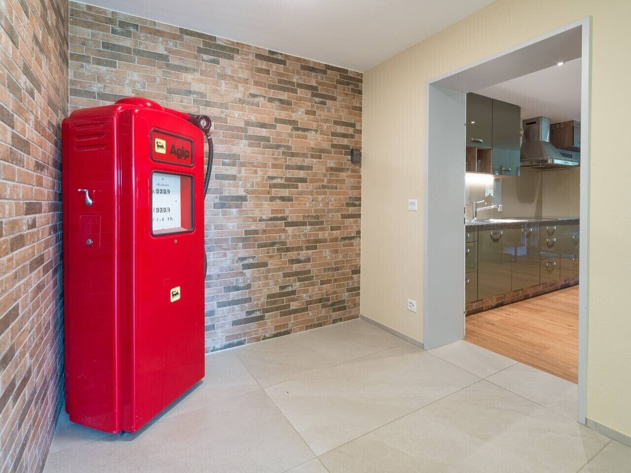 A red Agip refrigerator in a hallway with a brick wall and a glimpse of a kitchen area.