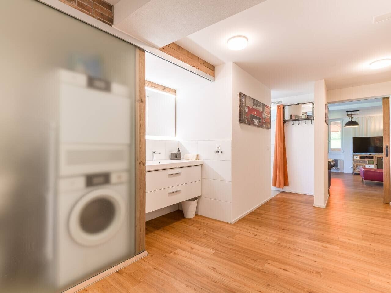 A modern apartment interior with a visible washing machine, a white bathroom with a sink, and a wooden floor.