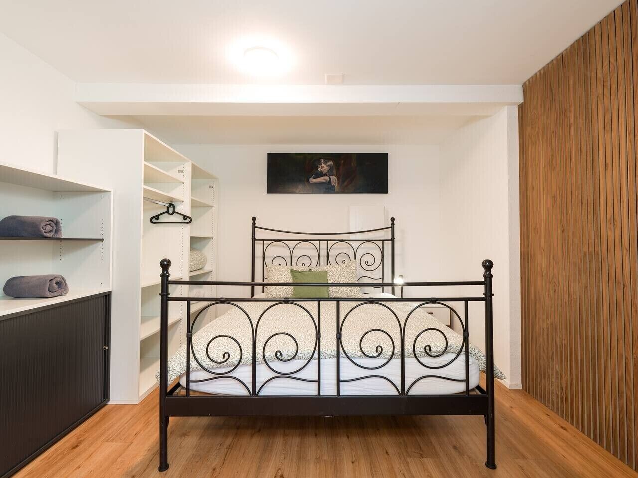 Una camera da letto con una struttura del letto in metallo, pavimento in legno, una parete bianca con un quadro e un armadio in legno.