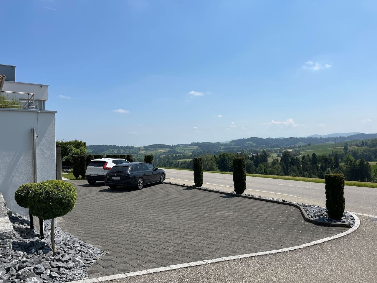 A parking area with two cars, a white and a black one, and a view of rolling hills in the background.