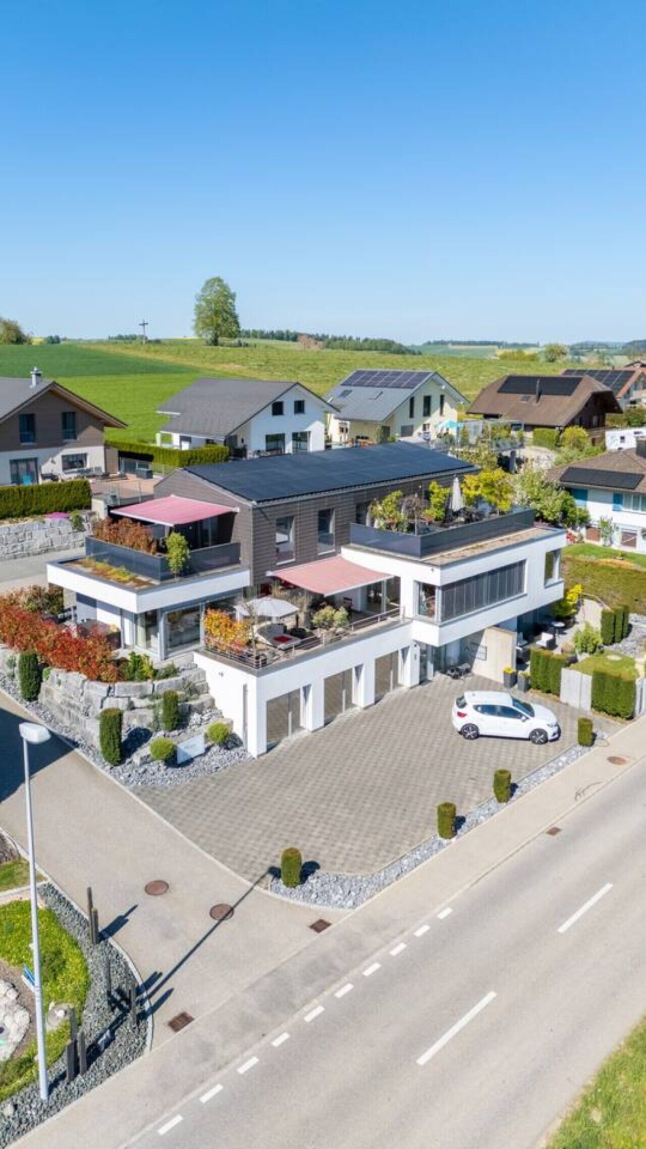 A modern multi-story residential building with a flat roof, solar panels, and a paved driveway.