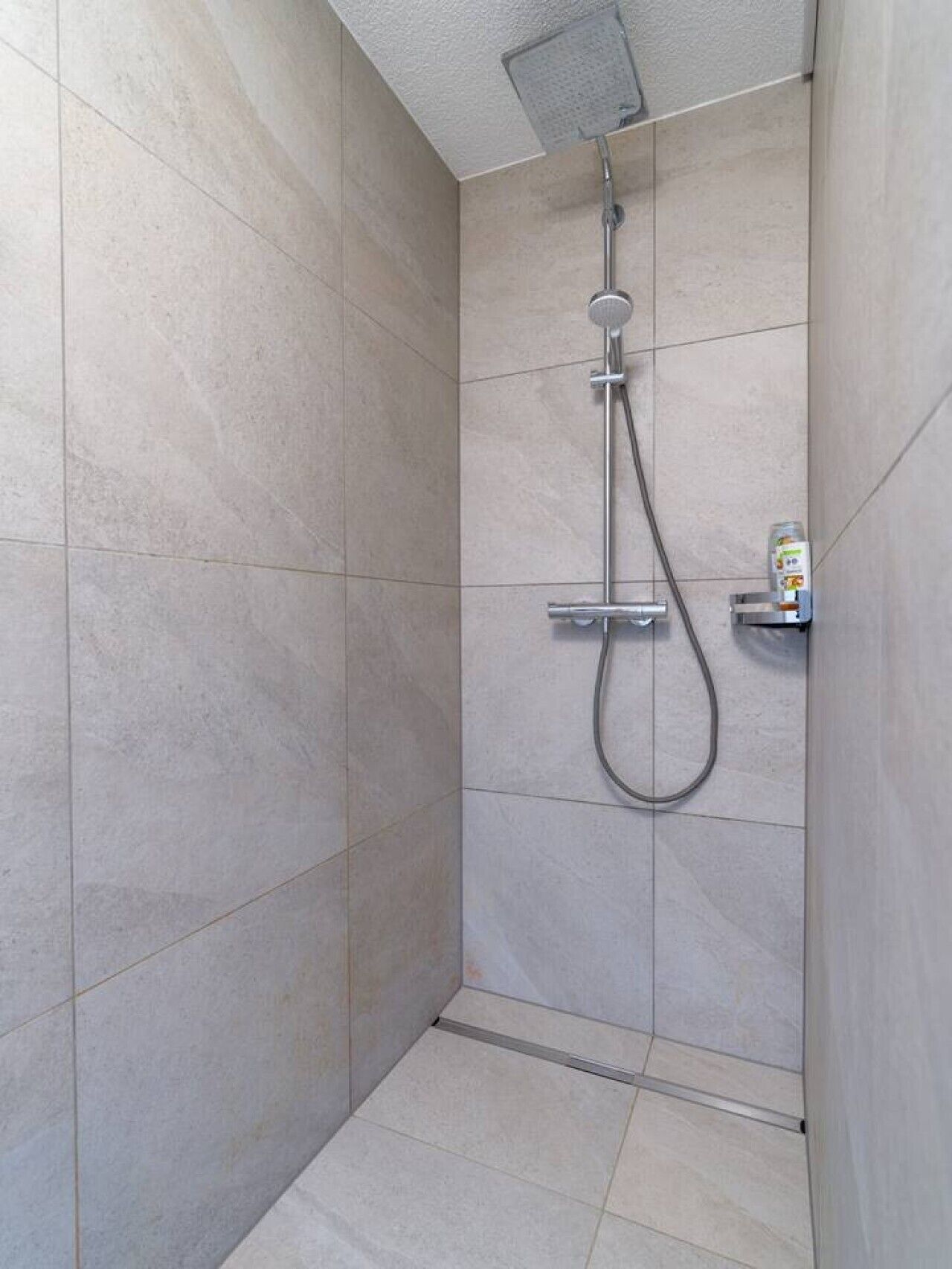 A shower with a handheld showerhead and a wall-mounted faucet, a small shelf with a bottle, and tiled walls and floor.