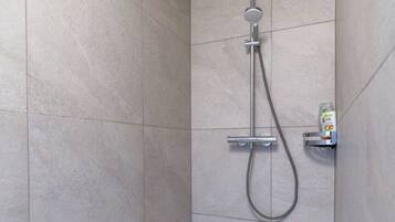 A shower with a handheld showerhead and a wall-mounted faucet, a small shelf with a bottle, and tiled walls and floor.