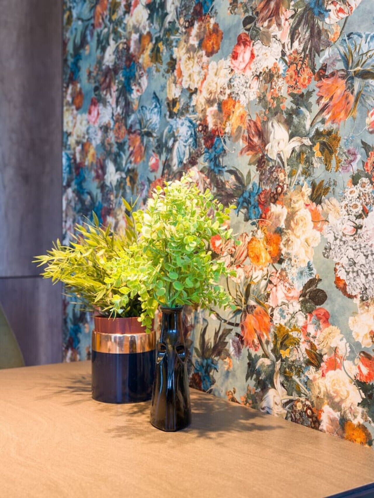 Une table sur laquelle sont posés deux vases contenant des plantes, le tout placé devant un papier peint à motifs floraux.