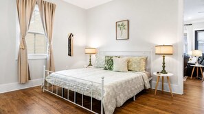 A bedroom with a bed, bedside lamps, a window with curtains, a wall-mounted picture, and a door leading to another room. - Heated Saltwater Pool - 3BR Home - Gulfport (Gulfport)