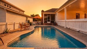 Ein Schwimmbecken mit Terrassenbereich, umgeben von einem Holzzaun, und ein Gebäude mit einer überdachten Veranda.