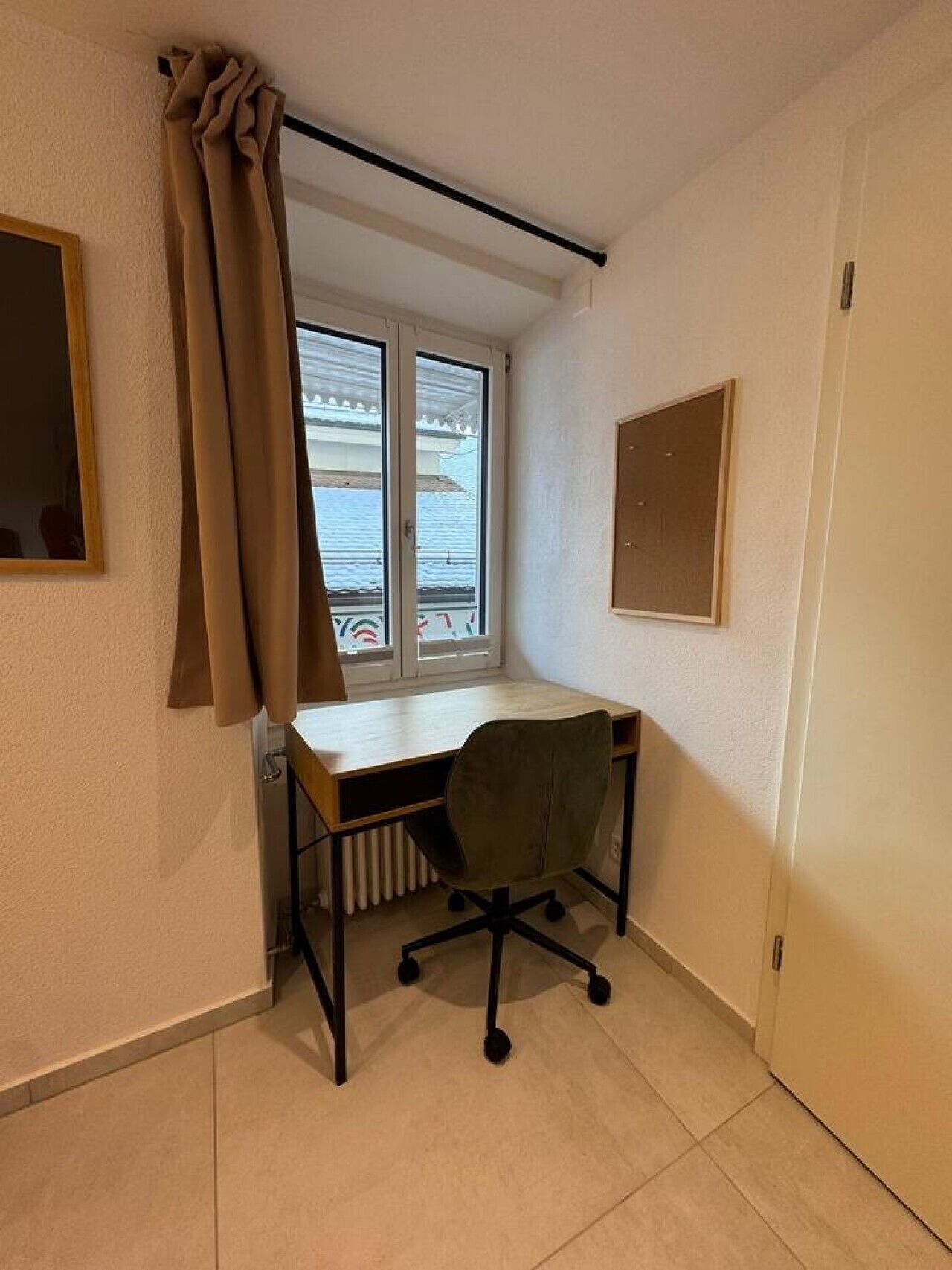 Ett litet rum med ett skrivbord, en stol, ett element, ett fönster, en gardin och en dörr.
