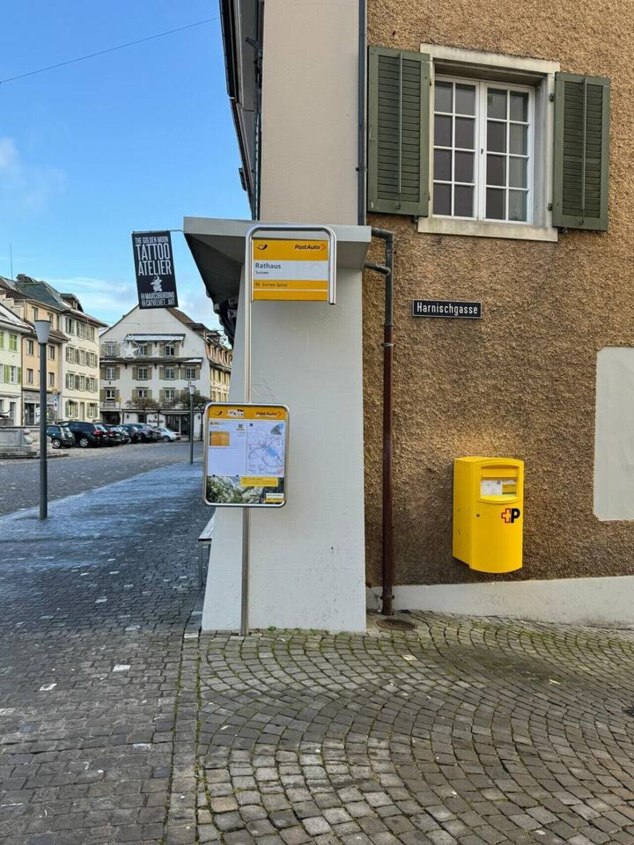 A street corner with a building featuring a PostAuto sign, a street sign for 'Harnischgasse,' and a yellow mailbox.