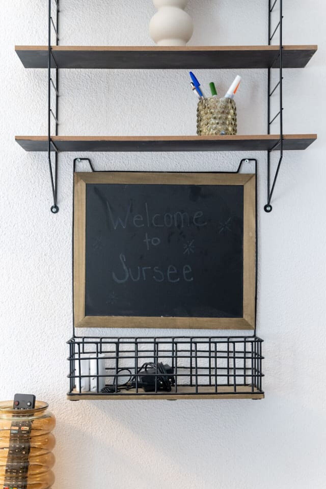 A blackboard with 'Welcome to Surrey' written on it, mounted on a wall with two shelves above and a wire basket below.