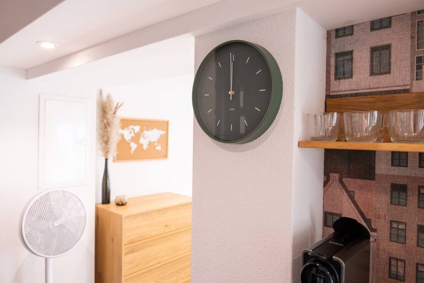 A room with a round wall clock, a world map, a fan, a wooden desk, and a shelf with glasses.