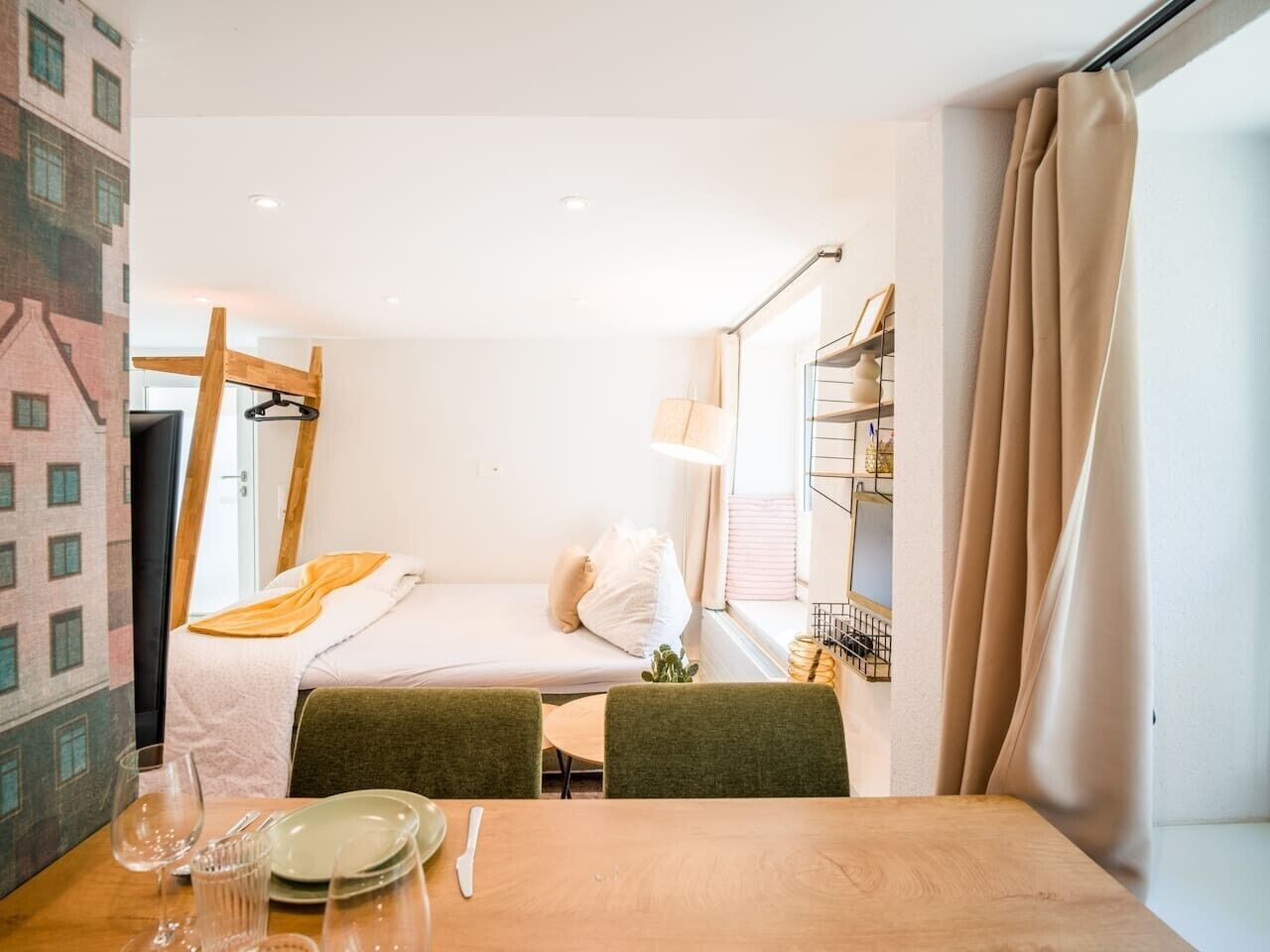 A compact hotel room with a single bed, a small table with a chair, a window with curtains, and a wall-mounted shelf.