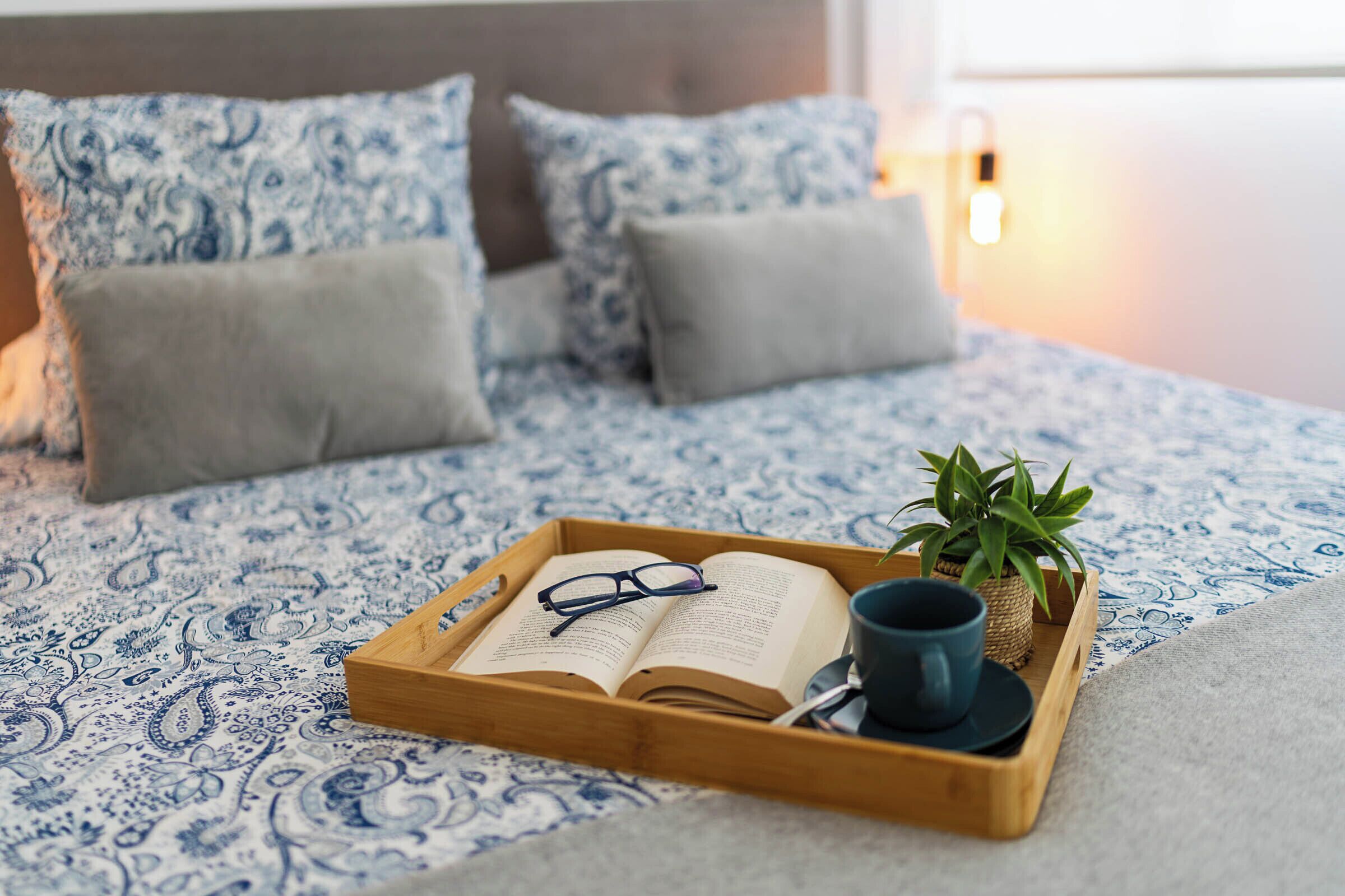 Una cama bien hecha con una colcha estampada, una bandeja con un libro abierto, vasos, una taza y una planta en maceta.