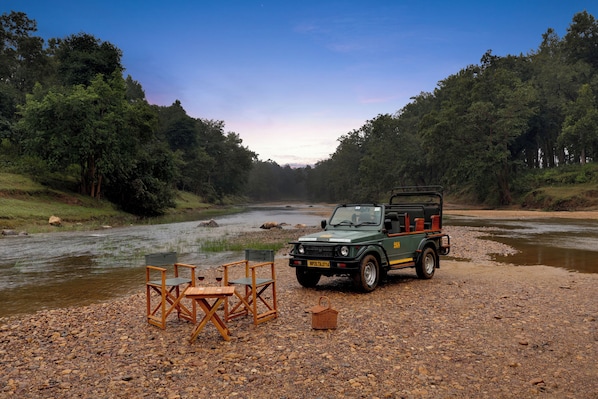 Un vehículo safari verde estacionado en un área de grava con un río al fondo y un bosque a cada lado.